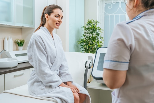 Beautiful smiling young woman in white bathrobe looking at cosmetologist in beauty clinic